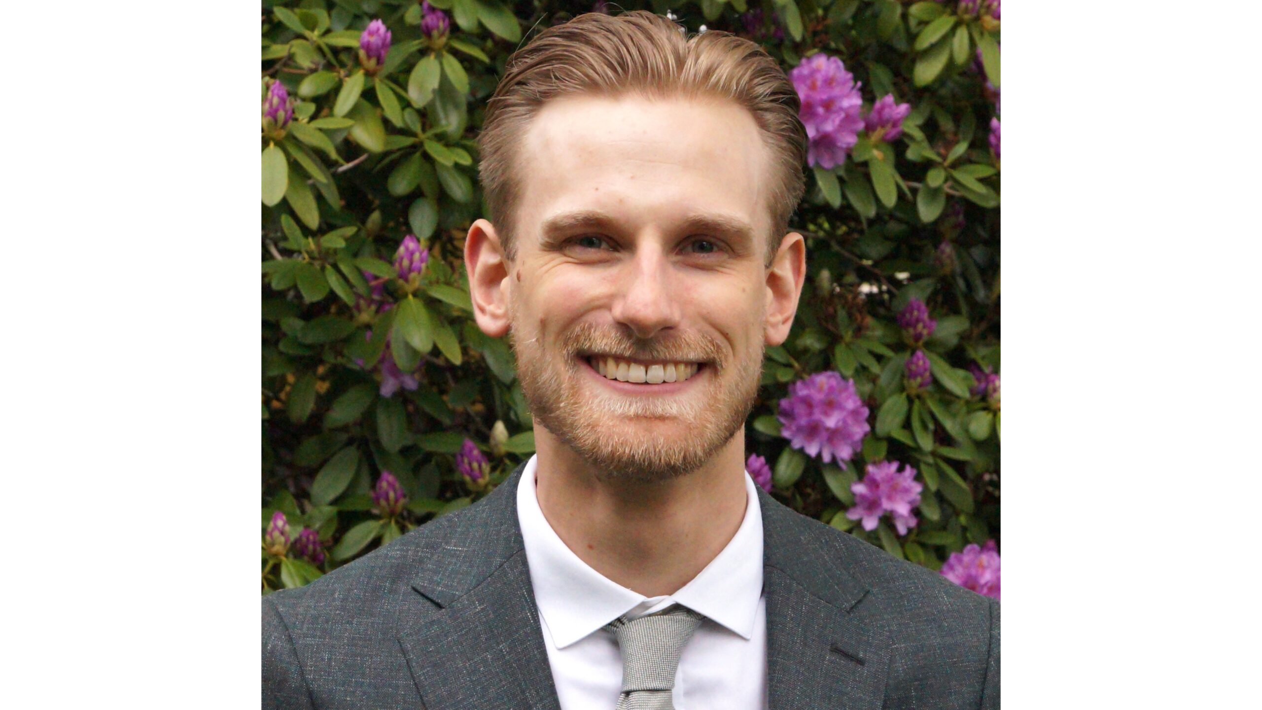 Johannes Widegren Young man smiling. Wearing a grey suite. Standing in front of bushes with purple flowers.