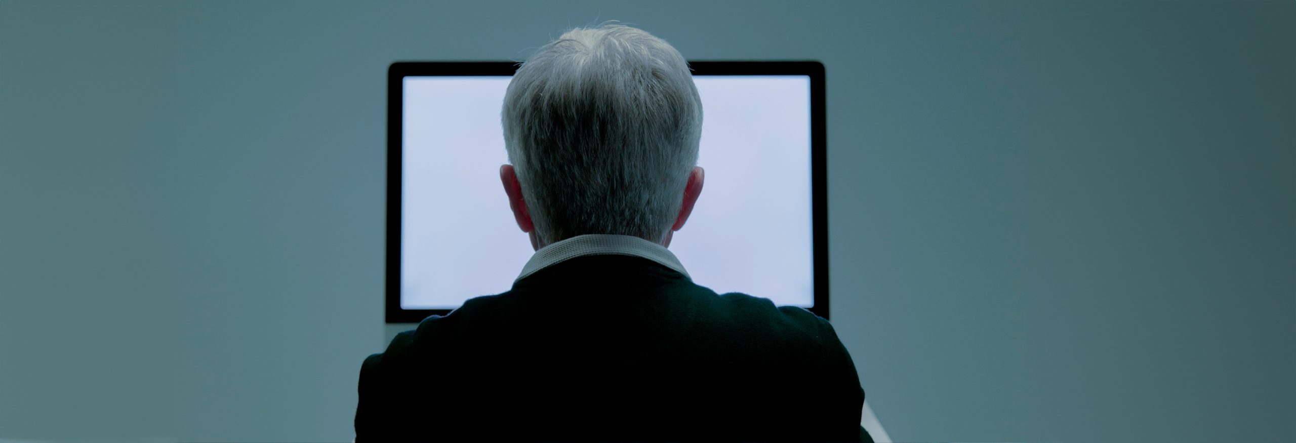 The back of an elderly man who is sitting in front of white monitor.
