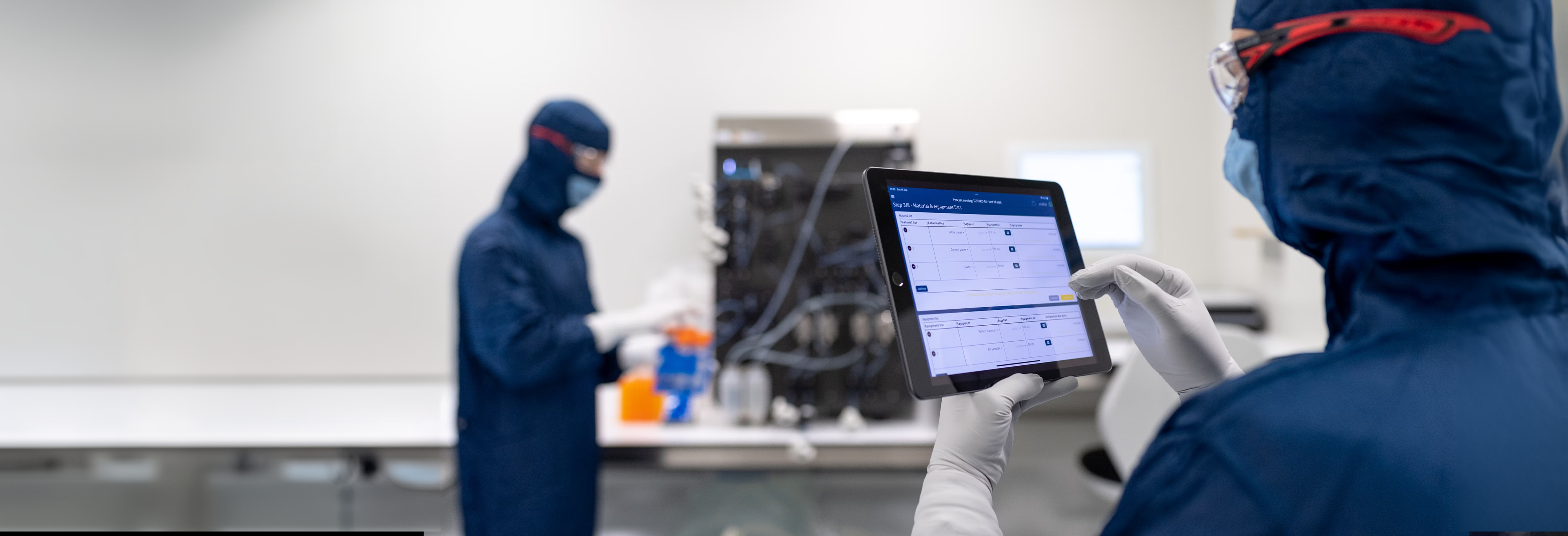 Two persons wearing blue full body covering clothes. Both interacting with digital devices in a white laboratory. Photo by Toon Lambrechts on Unsplash.