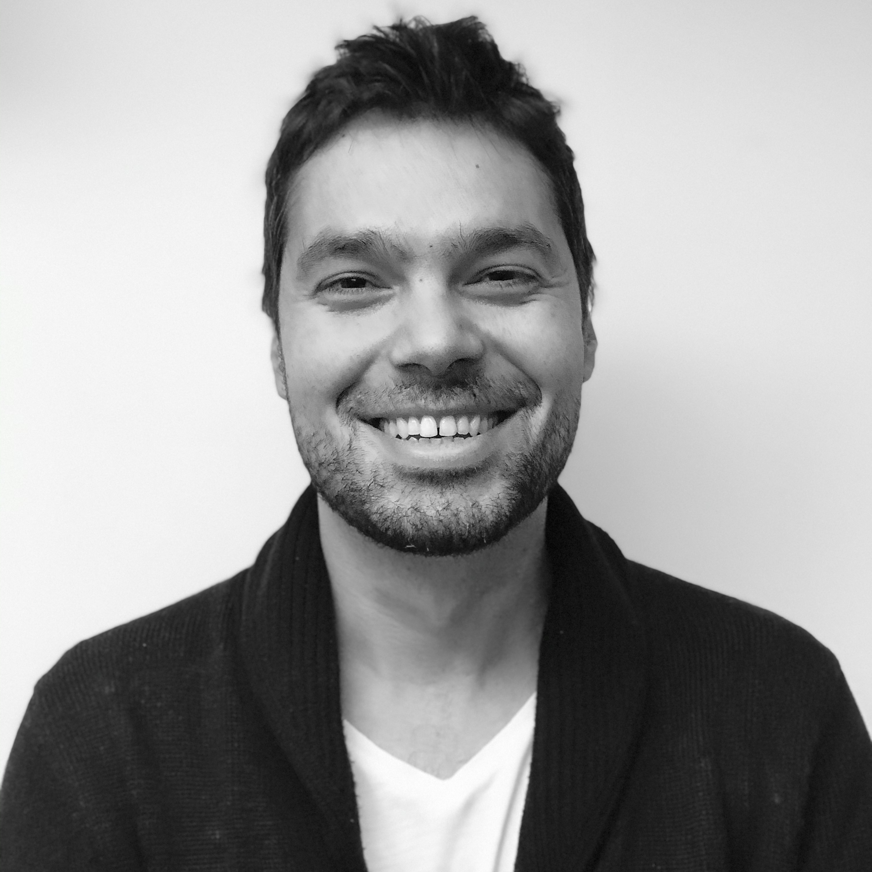 Black and white image of a dark haired man smiling for the camera. Portrait, chest up.